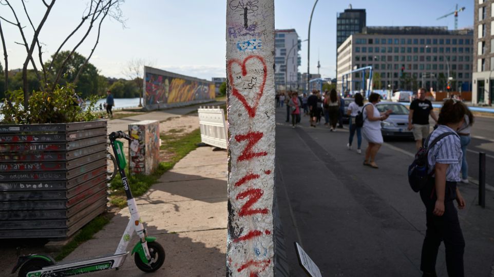 Ein bemaltes Segment der Berliner Mauer teilt die Ansicht: Links verläuft eine Uferpromenade mit E-Scooter, rechts ein Fußgängerweg mit Passanten