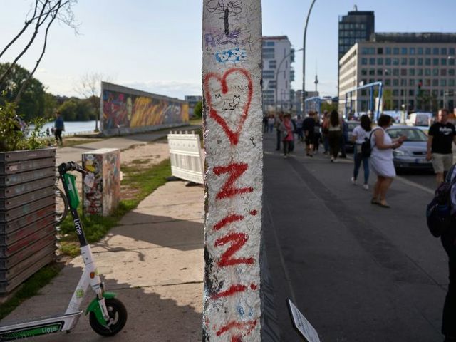 Ein bemaltes Segment der Berliner Mauer teilt die Ansicht: Links verläuft eine Uferpromenade mit E-Scooter, rechts ein Fußgängerweg mit Passanten