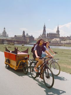 Zwei Fahrradfahrende mit Anhängern vor der Dresdener Altstadtkulisse.
