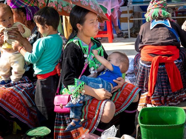 Mother breastfeeding her child at a market, other women and children sitting in the background