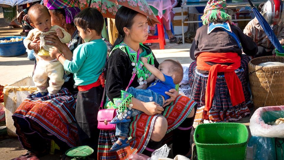 Mutter stillt ihr Kind auf einem Markt, im Hintergrund sitzen weitere Frauen und Kinder