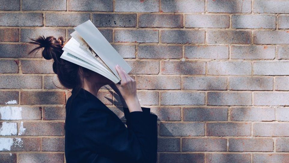 Junge Frau, die ihr Gesicht mit einem aufgeschlagenen Buch bedeckt.