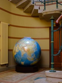 Großer Globus in einem alten Treppenhaus. Links von der Weltkugel ist eine Wandheizung zu sehen. Rechts führt eine Treppe mit rotem Teppich nach oben.