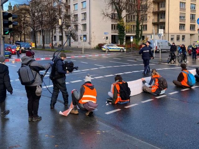 Sechs Menschen in Warnwesten blockieren eine Münchner Kreuzung. Sie werden von einem Fernsehteam interviewt.
