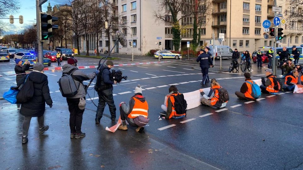 Blockadeaktion der "Letzten Generation" in München Sechs Menschen in Warnwesten blockieren eine Münchner Kreuzung. Sie werden von einem Fernsehteam interviewt.