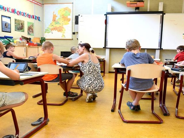 Grundschulklassenzimmer, Lehrerin sitzt in der Hocke neben Schüler und erklärt, alle anderen Schülerinnen und Schüler arbeiten mit gesenktem Kopf