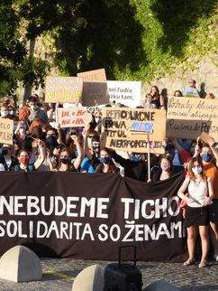 Demonstrierende auf der Straße vor einer Mauer und Bäumen. Viele halten Plakate in die Höhe, auf einem großen schwarzen Banner steht das Motto der Demonstration: »Nebudeme Ticho!« – »Wir werden nicht schweigen!« .