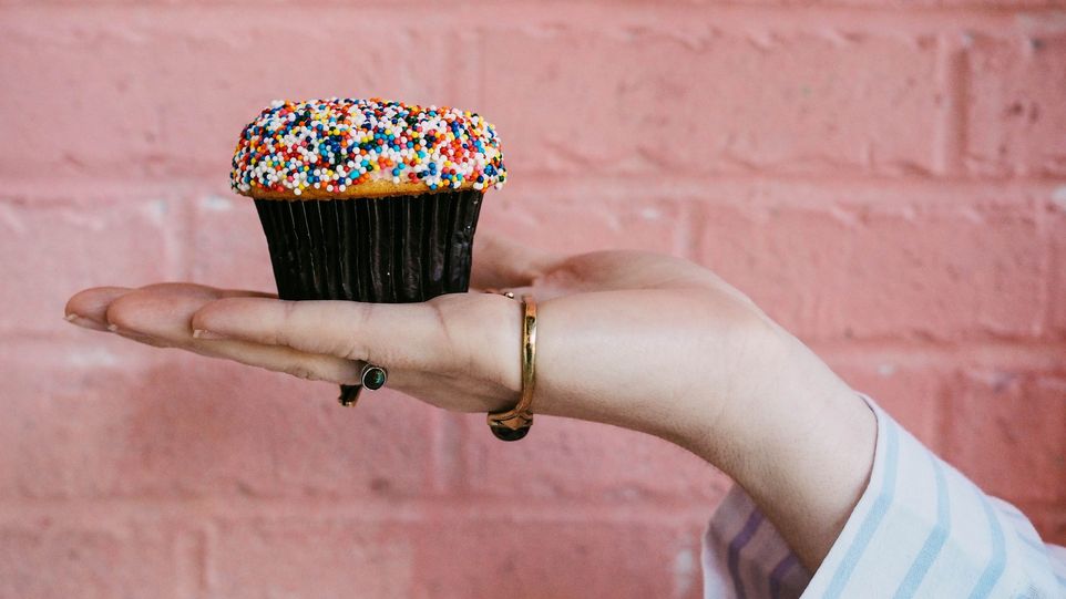 Eine Hand hält einen Muffin mit bunten Streuseln vor einer pinken Wand