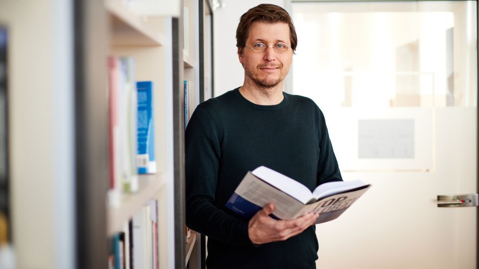 Moritz Schularick vor einem Bücherregal, ein aufgeschlagenes Buch in der Hand.