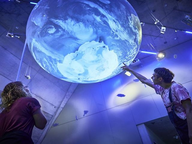 Besucherin und Besucher deuten auf großen blauen Globus, welcher von der Decke des Deutschen Museums hängt.