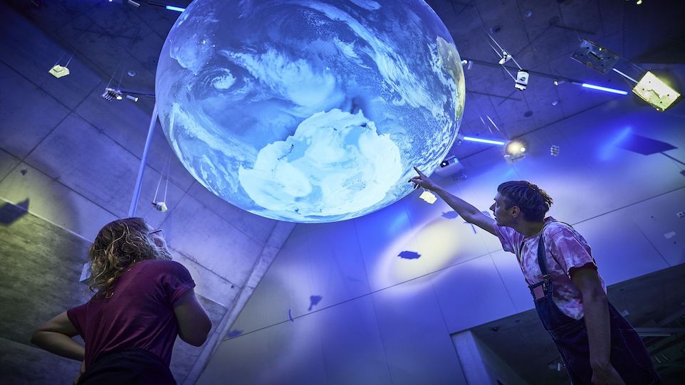 Besucherin und Besucher deuten auf großen blauen Globus, welcher von der Decke des Deutschen Museums hängt.