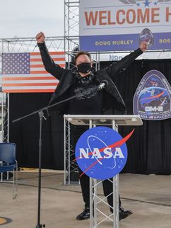 Auf einer Bühne feiert Elon Musk die Rückkehr der SpaceX-Astronauten Dough Hurley und Bob Behnke. Im Vordergrund das Logo der NASA, im Hintergrund die amerikanische Flagge und ein Flugzeug.