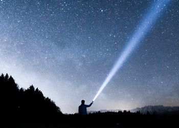 Das Bild zeigt einen klaren Sternenhimmel und die Silhouette eines Mannes mit Taschenlampe.
