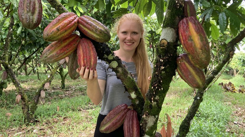 Das Foto zeigt Doktorandin Franziska Krause in einer Kakaoplantage in Nicaragua. Eine Frau steht hinter einem Kakaobaum und hält eine Kakaofrucht in der Hand