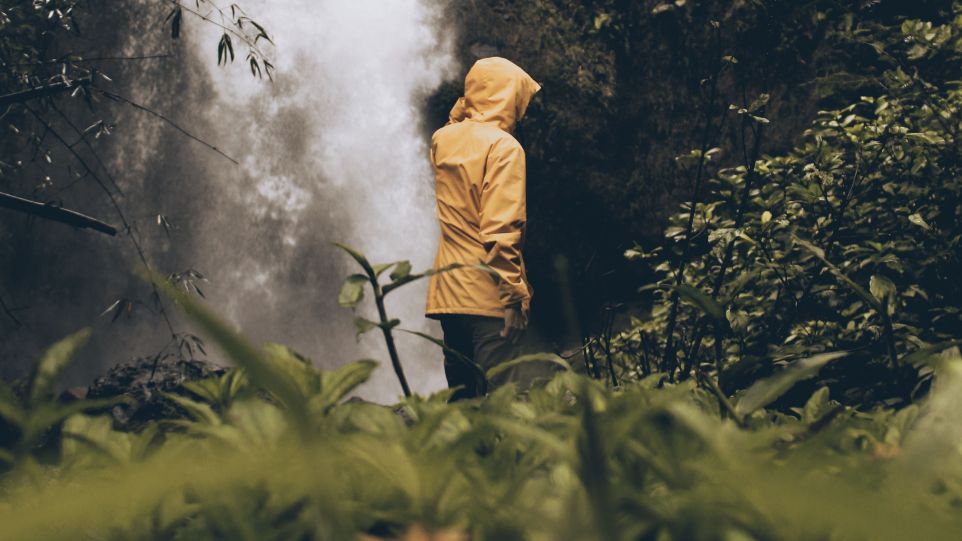 Rückenansicht einer Person im Regenmantel, vor einem im Wald gelegenen Wasserfall