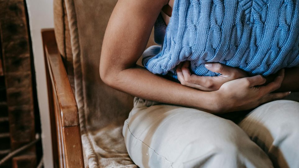 Eine gekrümmt sitzende Person in einem blauen Stricktop hält sich den Bauch