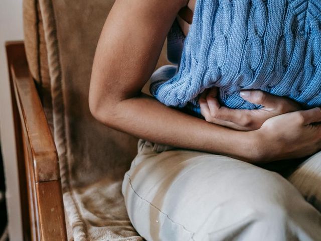 Eine gekrümmt sitzende Person in einem blauen Stricktop hält sich den Bauch