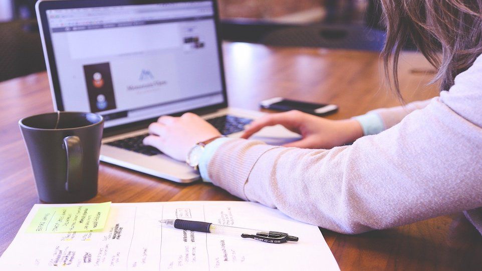 Frau arbeitet an Laptop, Kaffeetasse, Smartphone und Notizblock liegen daneben