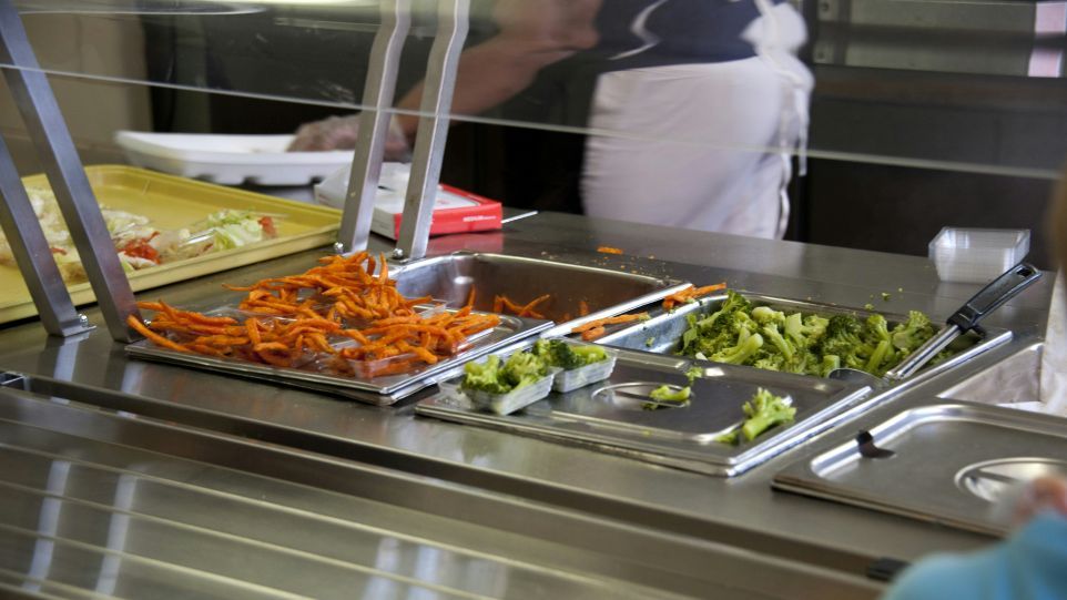 A salad bar display featuring stainless steel bowls filled with sweet potato fries and broccoli florets, with a person in white chef’s uniform standing behind it