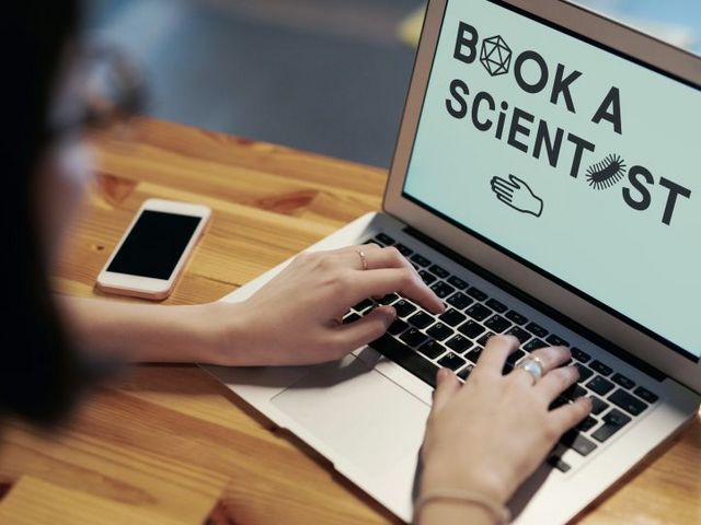 Frau am Laptop, auf dessen Bildschirm man die Zeile "Book a Scientist" lesen kann.