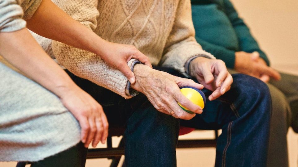 Ältere Person, die einen Stressball hält und von Pflegekraft unterstützt wird.