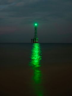 Ein turmähnliches Metallgerüst mit grünem Licht ragt aus dem Meer. Das Licht spiegelt sich als langer grüner Streifen auf der ruhigen Wasseroberfläche, während der Nachthimmel dunkel und wolkenverhangen wirkt.