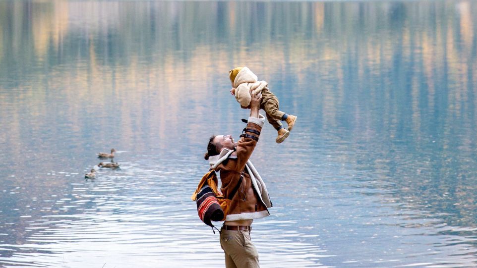 Ein Mann hebt ein Kleinkind hoch, im Hintergrund ein See mit Enten