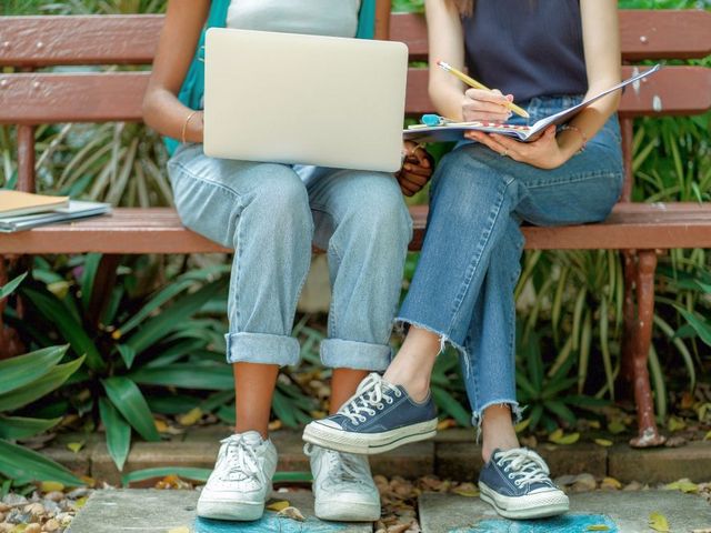Zwei Schülerinnen auf einer Parkbank, die eine hält ein Laptop auf dem Schoß, die andere Heft und Stift.