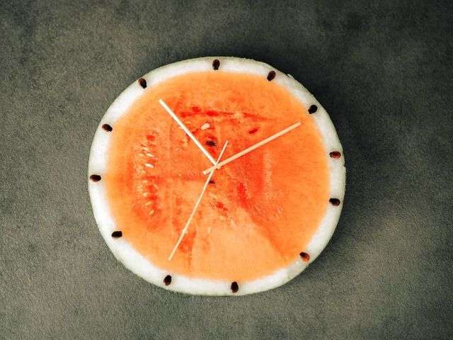 Draufsicht einer halbierten Melone, Kerne und Holzstäbchen symbolisieren Uhrzeiger und Minuten