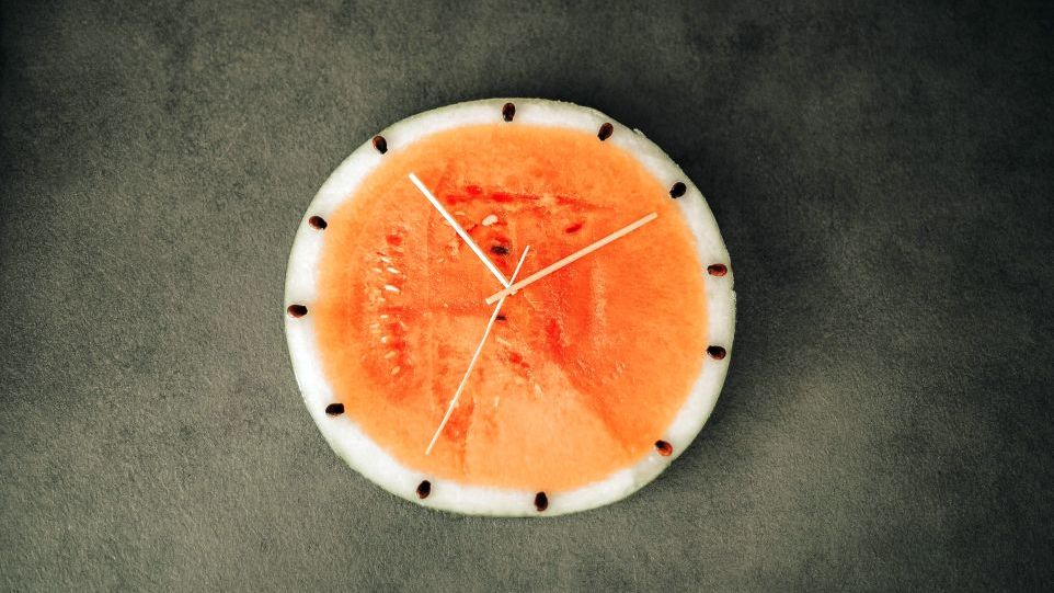 Draufsicht einer halbierten Melone, Kerne und Holzstäbchen symbolisieren Uhrzeiger und Minuten