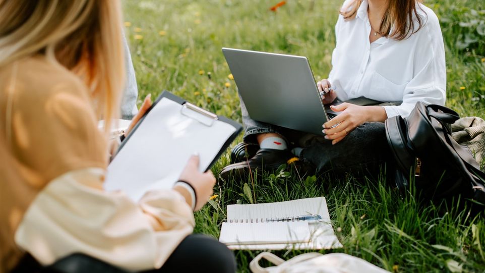 Zwei junge Frauen sitzen mit Schreibblock und Laptop im Gras