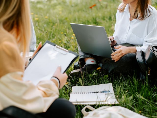 Zwei junge Frauen sitzen mit Schreibblock und Laptop im Gras