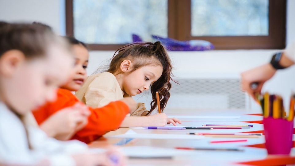 Drei Mädchen sitzen in einem Klassenzimmer, auf den Tischen vor ihnen liegen Stifte und Papier.