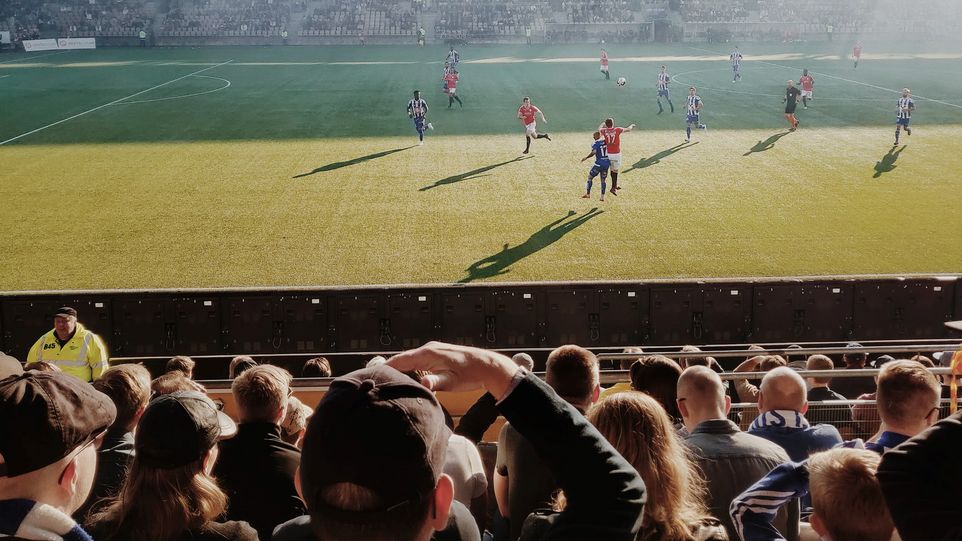 Fußballtribüne mit Blick auf das Spielfeld, auf welchem ein Spiel im Sonnenlicht stattfindet