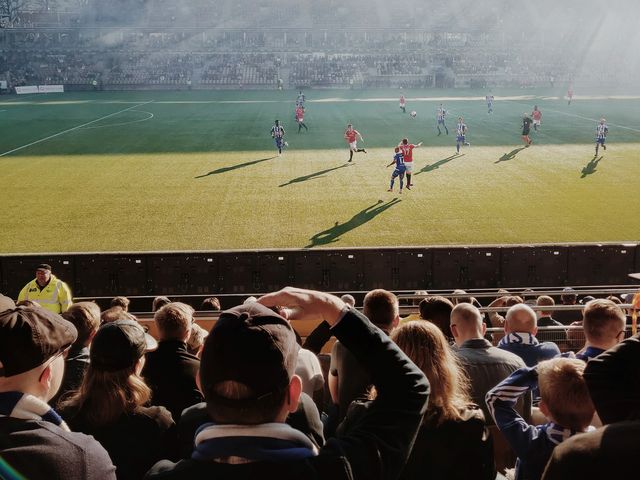 Fußballtribüne mit Blick auf das Spielfeld, auf welchem ein Spiel im Sonnenlicht stattfindet