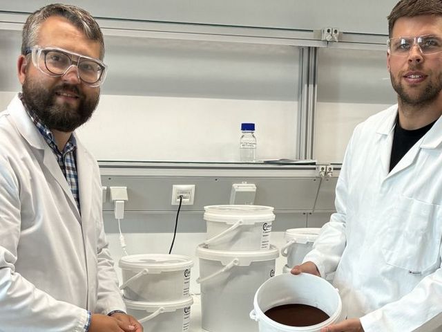 Zwei Männer mit Kitteln und Schutzbrillen präsentieren einen mit braunem Material gefüllten Eimer