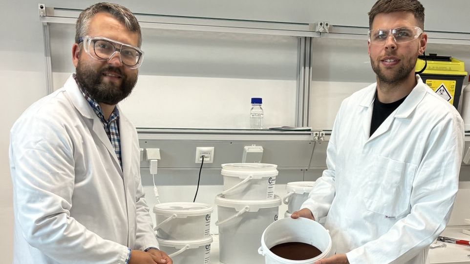 Ein Teil des synthetisierten Materials wird von Dr. Christoph Wulf (links) und Dr. Simon Haida präsentiert. Zwei Männer mit Kitteln und Schutzbrillen präsentieren einen mit braunem Material gefüllten Eimer