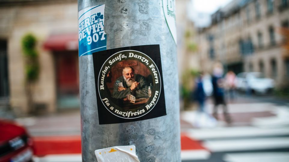 Aufkleber auf Straßenpfahl mit der Aufschrift: Raucha, Saufa, Danzn, Feiern - fia a nazifreies Bayern.